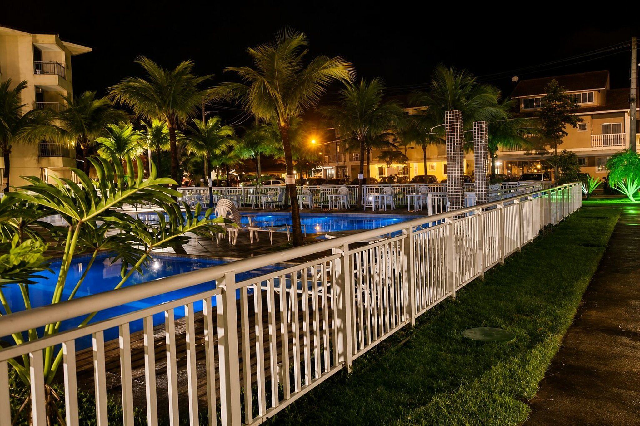 Vista noturna de uma área de piscina de hotel, com cerca, palmeiras e edifícios ao fundo.