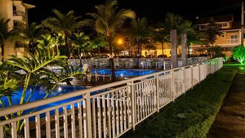 Vista noturna de uma área de piscina de hotel, com cerca, palmeiras e edifícios ao fundo.