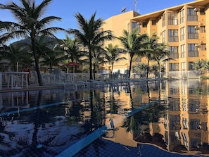 A swimming pool with clear water, surrounded by palm trees and a building with balconies. - Reserva do Sahy, Condado (Mangaratiba)