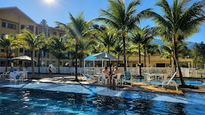 Ein Poolbereich mit Liegestühlen und Sonnenschirmen, umgeben von Palmen und einem Gebäude mit zahlreichen Fenstern.