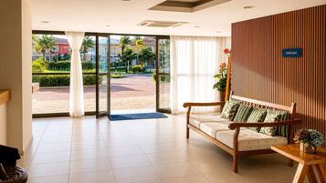 Um saguão de hotel com balcão de recepção de madeira, um sofá com almofadas estampadas e uma grande porta de vidro que dá para área externa com vista para palmeiras e prédios.