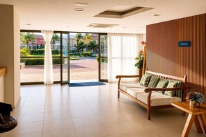 Eine Hotelhalle mit einem hölzernen Empfangsbereich, einer Couch mit gemusterten Kissen und einer großen Glasdoor, die zu einem Außenbereich mit Blick auf Palmen und Gebäude führt.