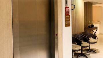 Uma área de recepção de hotel com elevador fechado, balcão de recepção e área de estar com cadeiras.