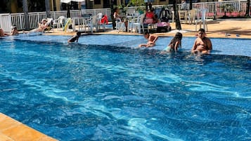Piscina com pessoas na água, rodeada por espreguiçadeiras e guarda-sóis.