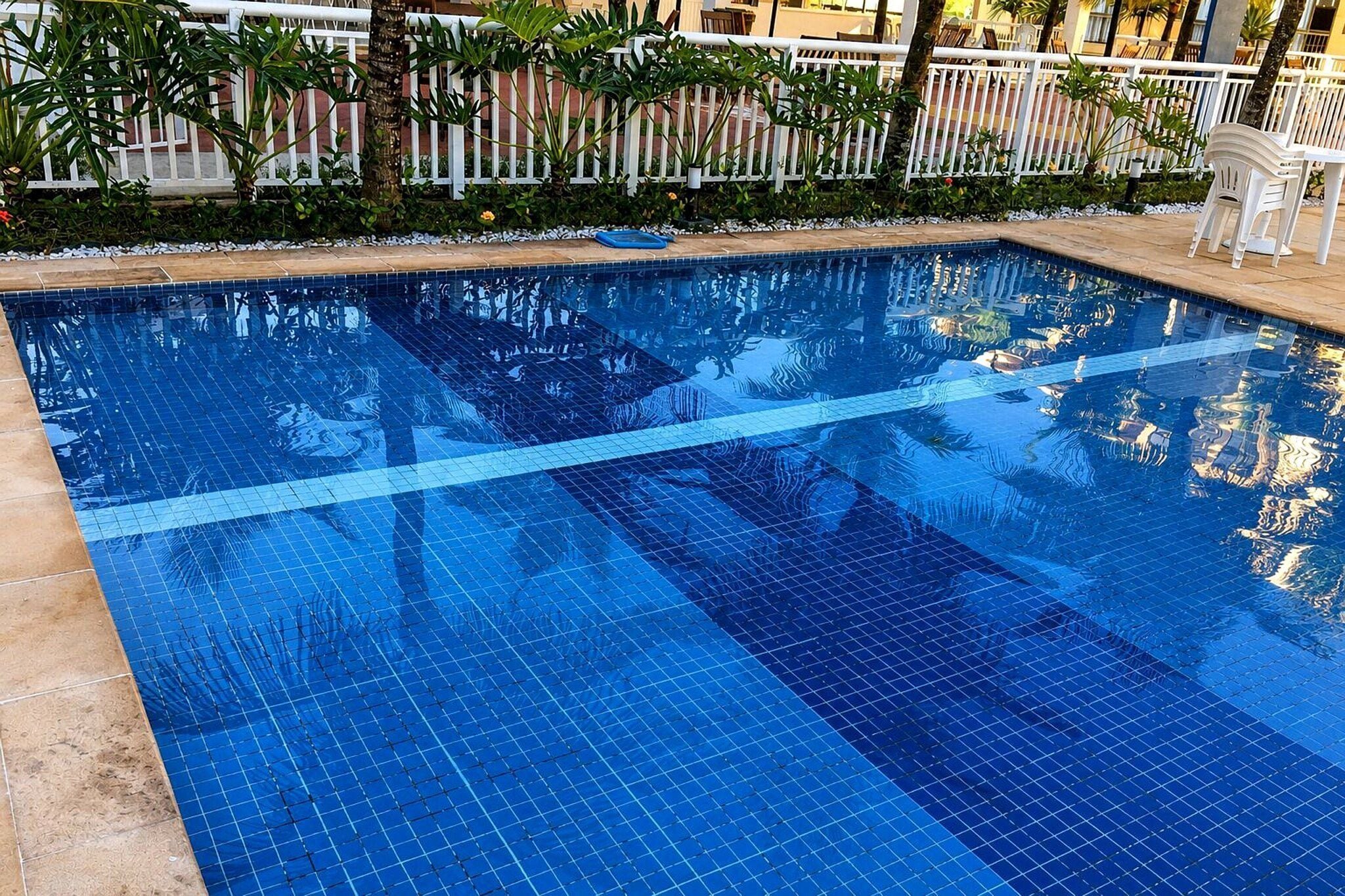 Piscina azul límpida com borda em azulejo e cerca branca ao fundo.