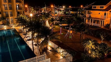 Vista noturna de um resort com piscina, palmeiras e edifícios iluminados.