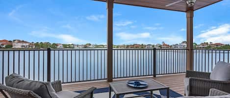 A covered outdoor deck with wicker furniture, a table, and a view of a lake with houses in the background.