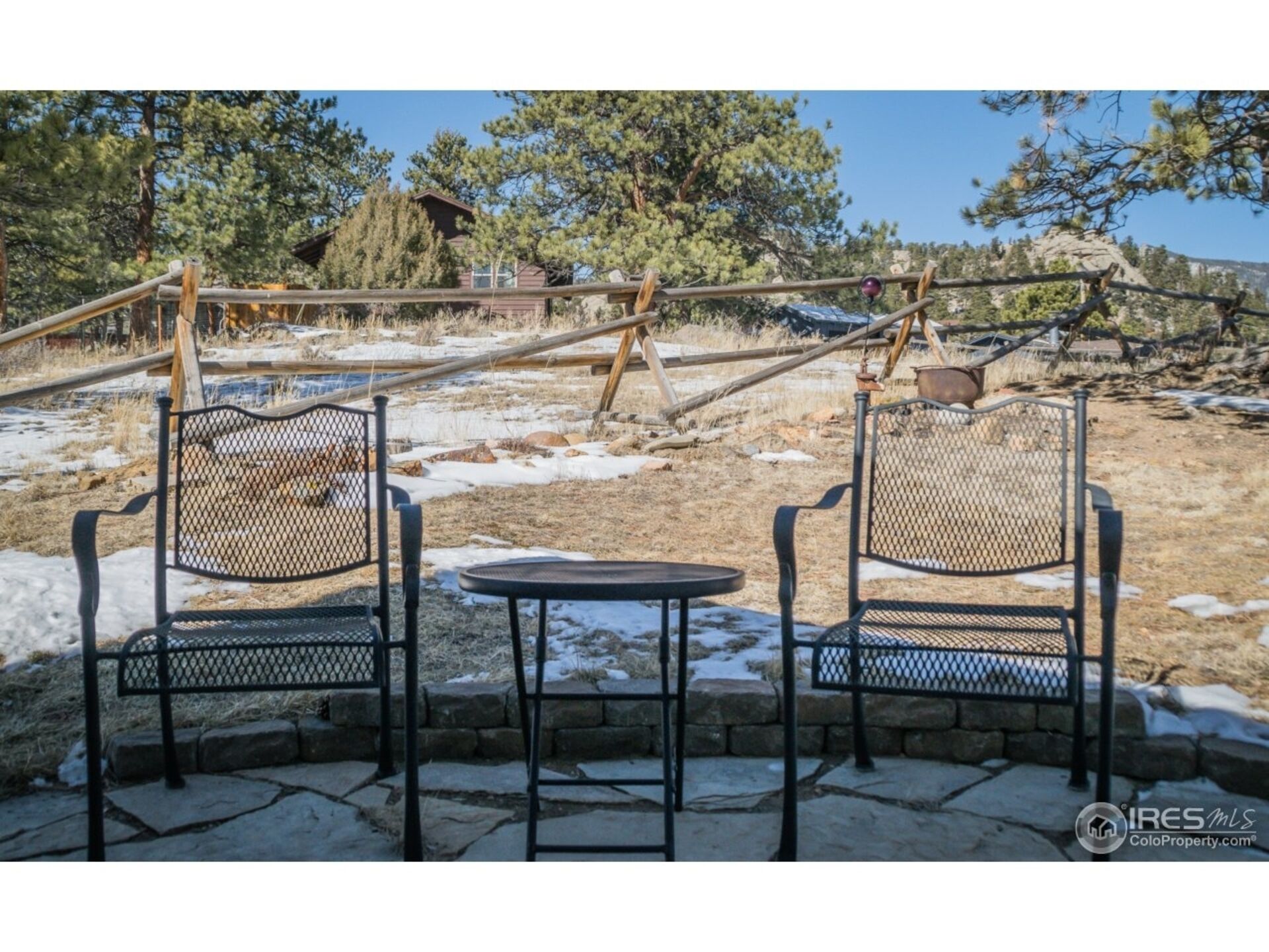 Two metal chairs and a small table in a stone patio area with a view of a rustic cabin and trees.