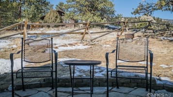 Two metal chairs and a small table in a stone patio area with a view of a rustic cabin and trees.