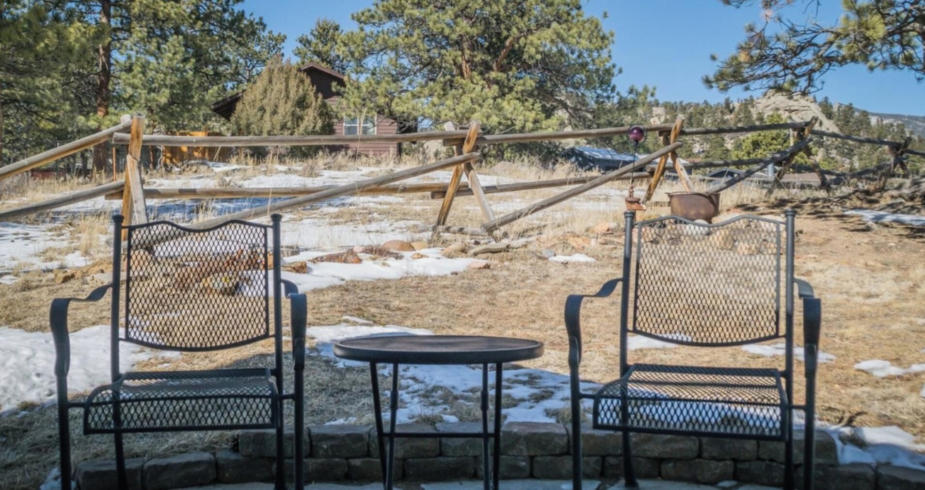 Two metal chairs and a small table in a stone patio area with a view of a rustic cabin and trees.