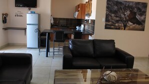 A compact living space with a kitchenette, a black leather sofa, a glass coffee table, and a painting of a tree on the wall. - Eldorado Lodge CC (Kuruman)