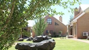 A courtyard with large rocks, a brick building, and a tree. - Eldorado Lodge CC (Kuruman)