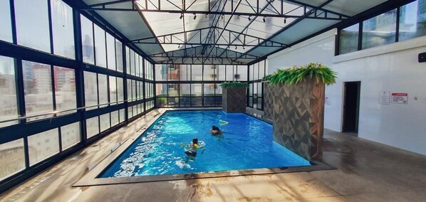 Una piscina interior con paredes de vidrio transparente, piso de baldosas y estructura de techo metálica.