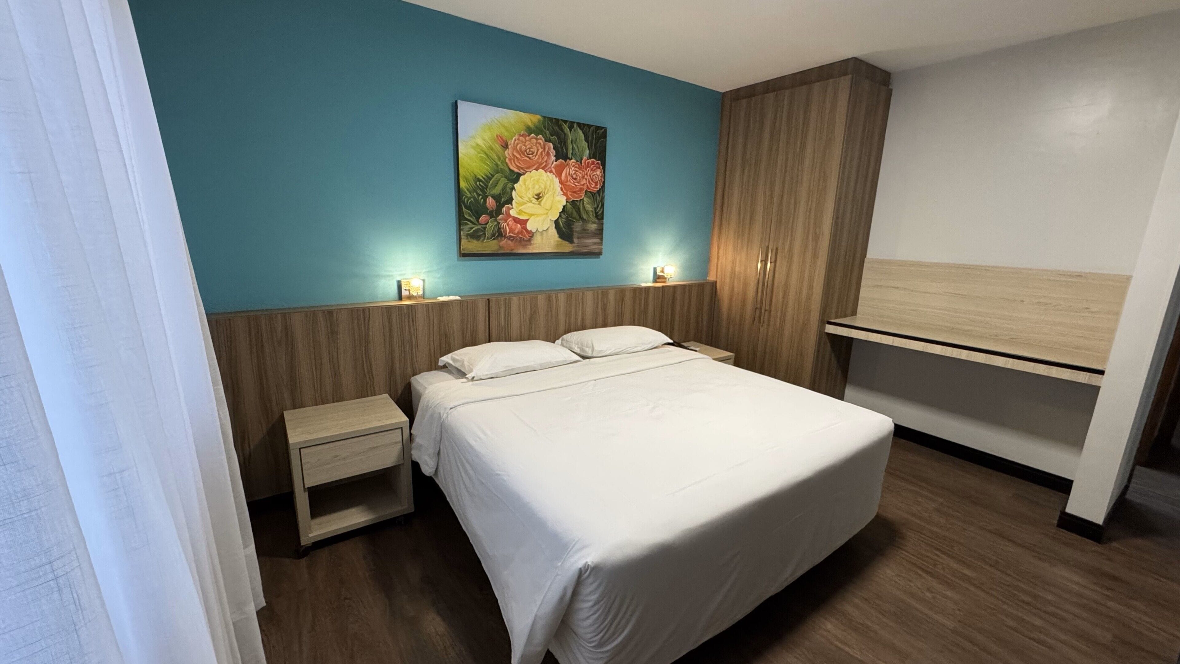 A hotel room with a bed, bedside tables, a painting on the wall, and a wardrobe.