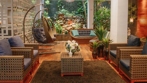 A cozy indoor seating area with wicker furniture, a small table with a vase of flowers, and a backdrop of a stone wall and greenery. - Hotel das Américas (Balneário Camboriú)