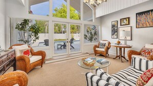 Ein geräumiges Wohnzimmer mit großen Fenstern, Korbmöbeln, einem Glastisch und einer modernen Pendelleuchte.