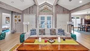 Ein geräumiges Wohnzimmer mit hohen Decken, Holzvertäfelung, einem großen Sofa mit gemusterten Kissen, einem Holztisch mit Kerzen und einem Glasbehälter sowie ein Essbereich, der durch eine geöffnete Tür sichtbar ist.
