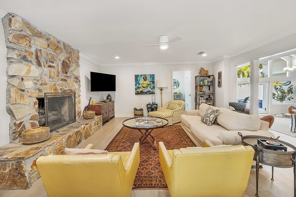 A living room with a stone fireplace, a sofa, yellow chairs, a coffee table, and a bookshelf. - GOOD TIDES - Steps from East Beach AND has a large private pool! (Saint Simons Island)