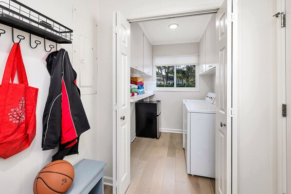 A narrow hallway leading to a laundry area with a washing machine and dryer, a small window with a view of trees, and a coat rack with a red bag and a black jacket.