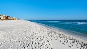 A wide sandy beach with footprints leading to the water, a clear blue sky, and buildings in the background. - Ocean view, walk to the beach, pool,  military/first responder discount 13A (Destin)