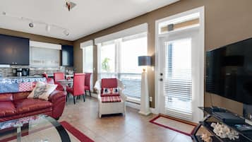 Una sala moderna con un sofá de cuero rojo, una mesa de centro de vidrio y un comedor con sillas rojas. Se observa una cocina con armarios oscuros y un ventanal grande.