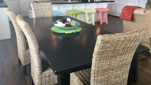 Un comedor con una mesa negra rodeada de sillas de mimbre, un plato con piñas y un bol, y una cocina al fondo.