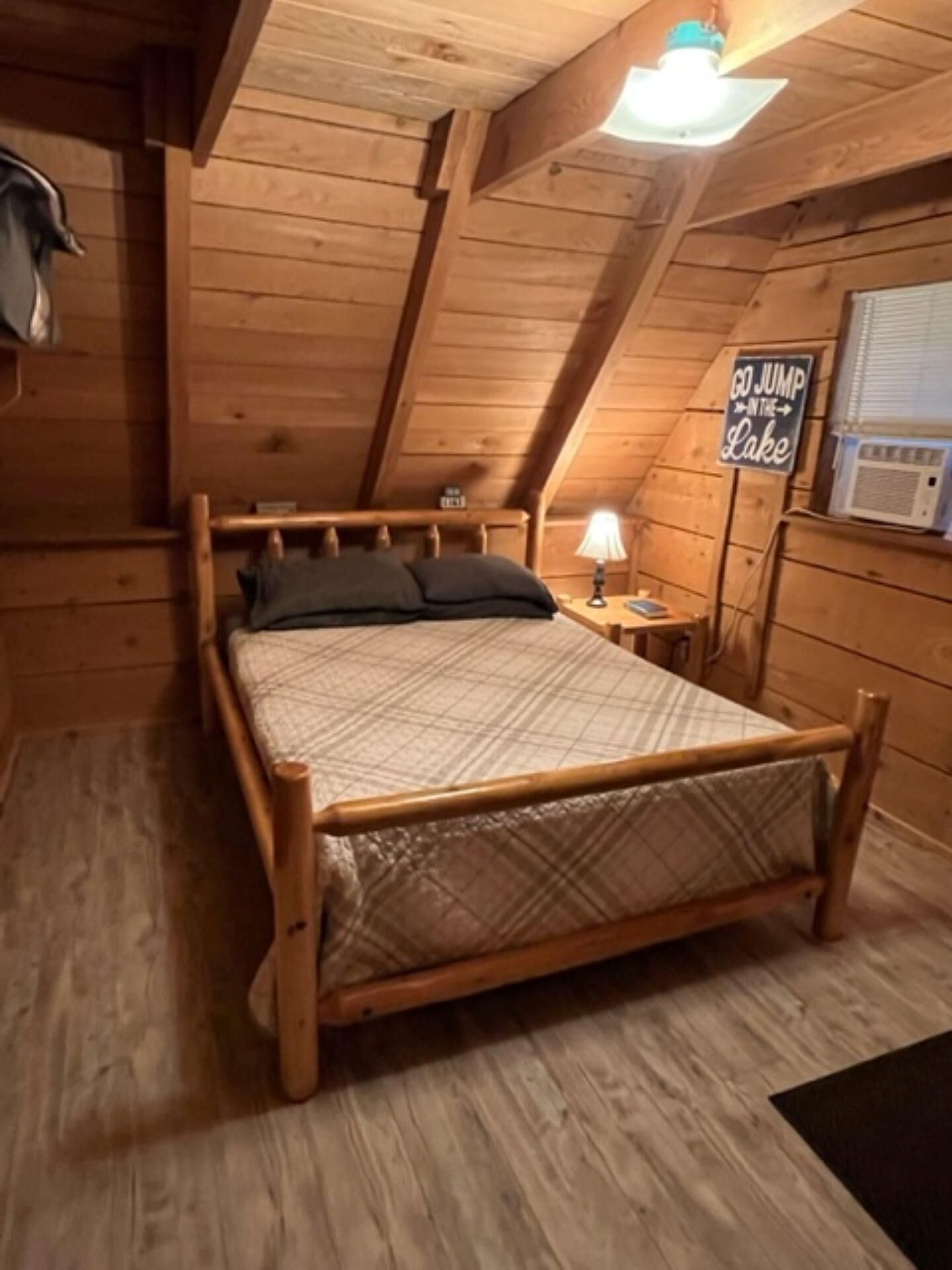 A wooden cabin bedroom with a bed, a nightstand, a lamp, and a sign that says "GO JUMP IN THE LAKE".