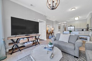 A modern living room with a flat-screen TV, a grey sofa, a coffee table, and a kitchen in the background. - Overlooking 30A!  Great Location! Easy Beach & Pool Access! (Seacrest Beach)