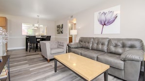 Una sala de estar con un sofá gris, una mesa de centro de madera, un sillón gris y una zona de comedor con una mesa y sillas.