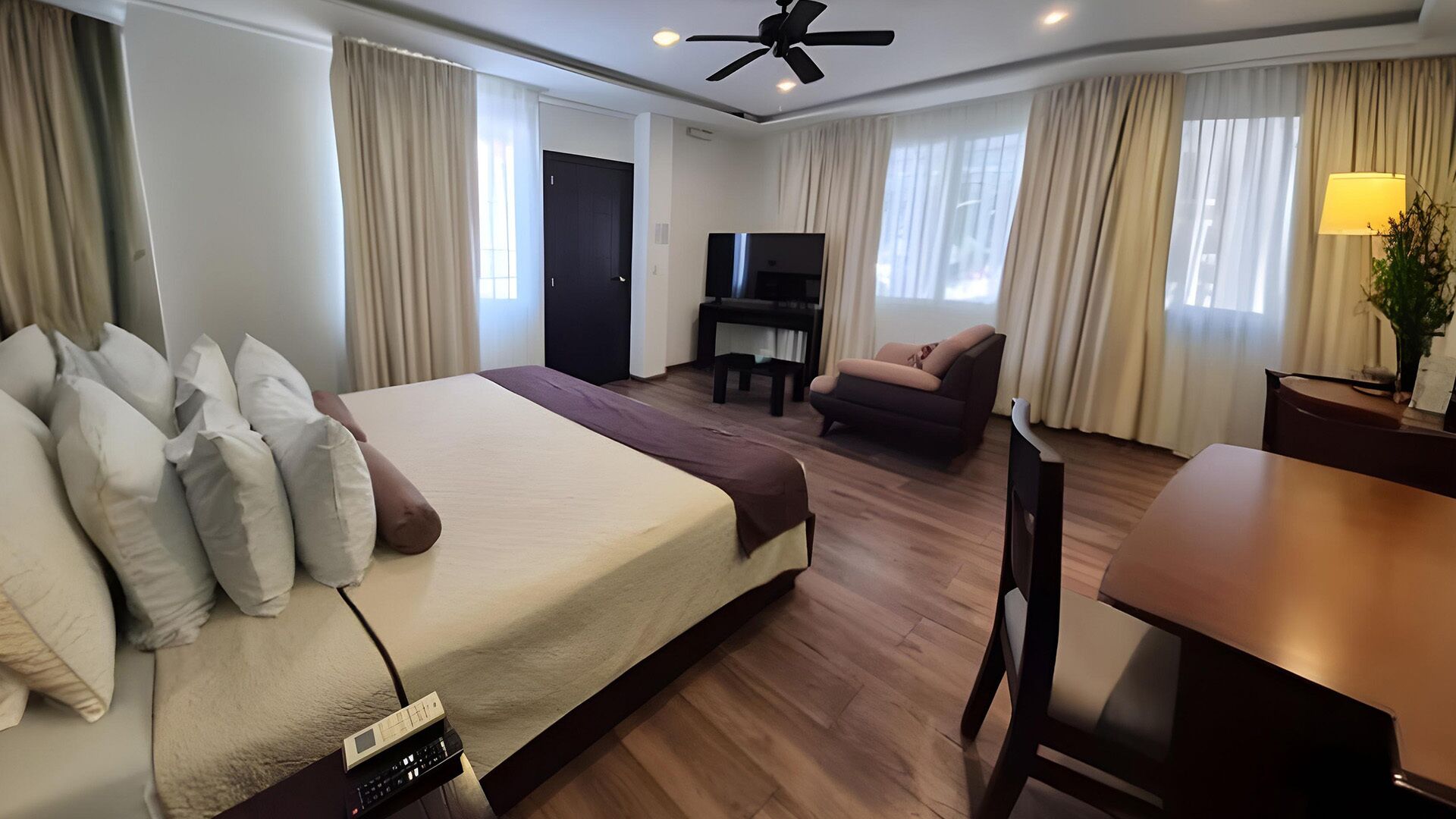 A hotel room with a large bed, a desk, a chair, a television, and a ceiling fan.