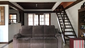 Ein Wohnzimmer mit einem großen grauen Sofa, einer Holztreppe und einem durchgegangenen Tür erkennbaren Küchenbereich.