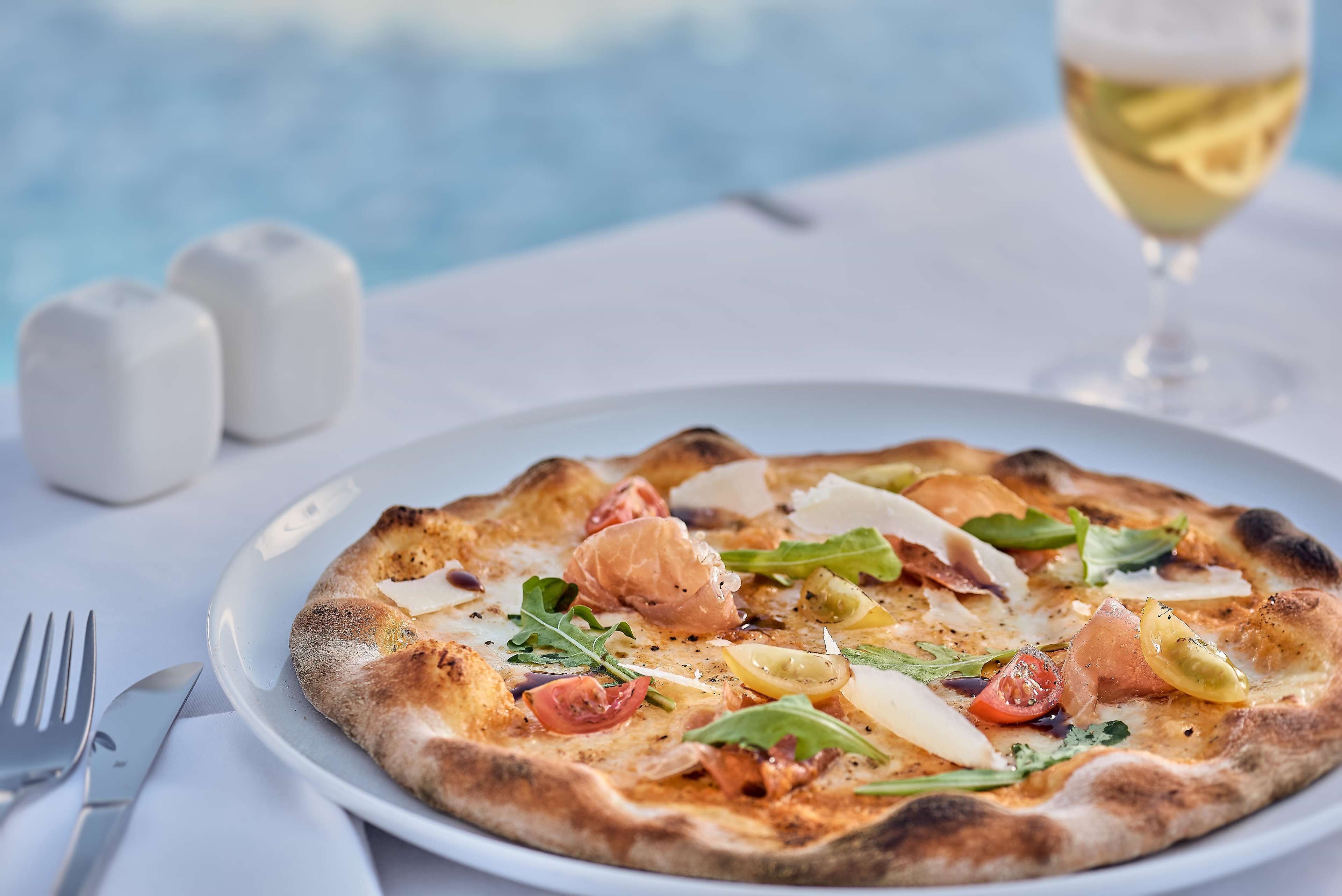 Una pizza con diversos ingredientes sobre un plato blanco, colocada sobre una mesa con una copa de vino blanco y saleros y pimenteros al fondo.
