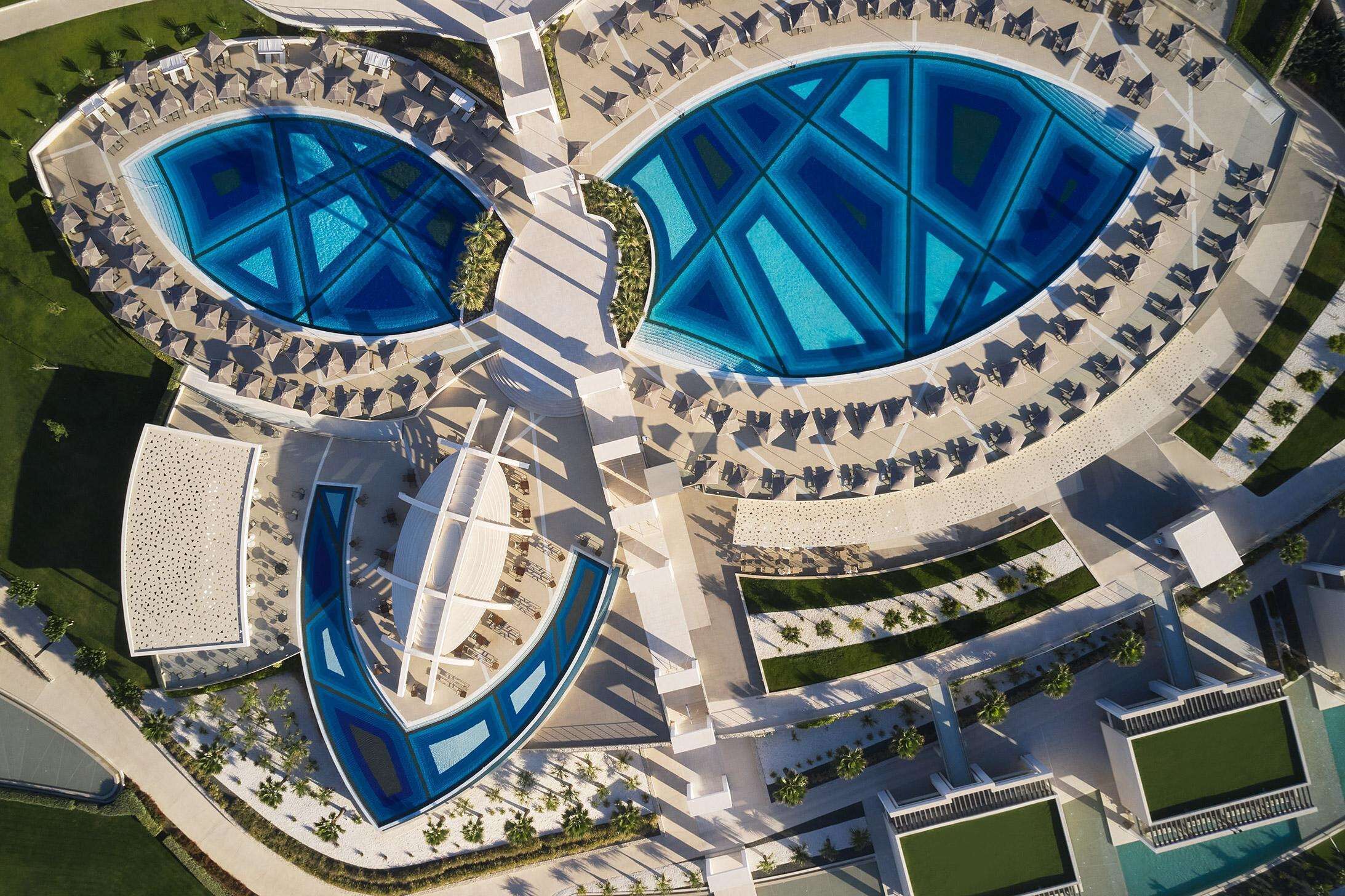 Vista aérea de un hotel con dos piscinas grandes, sillas de descanso y un área de estar cubierta.