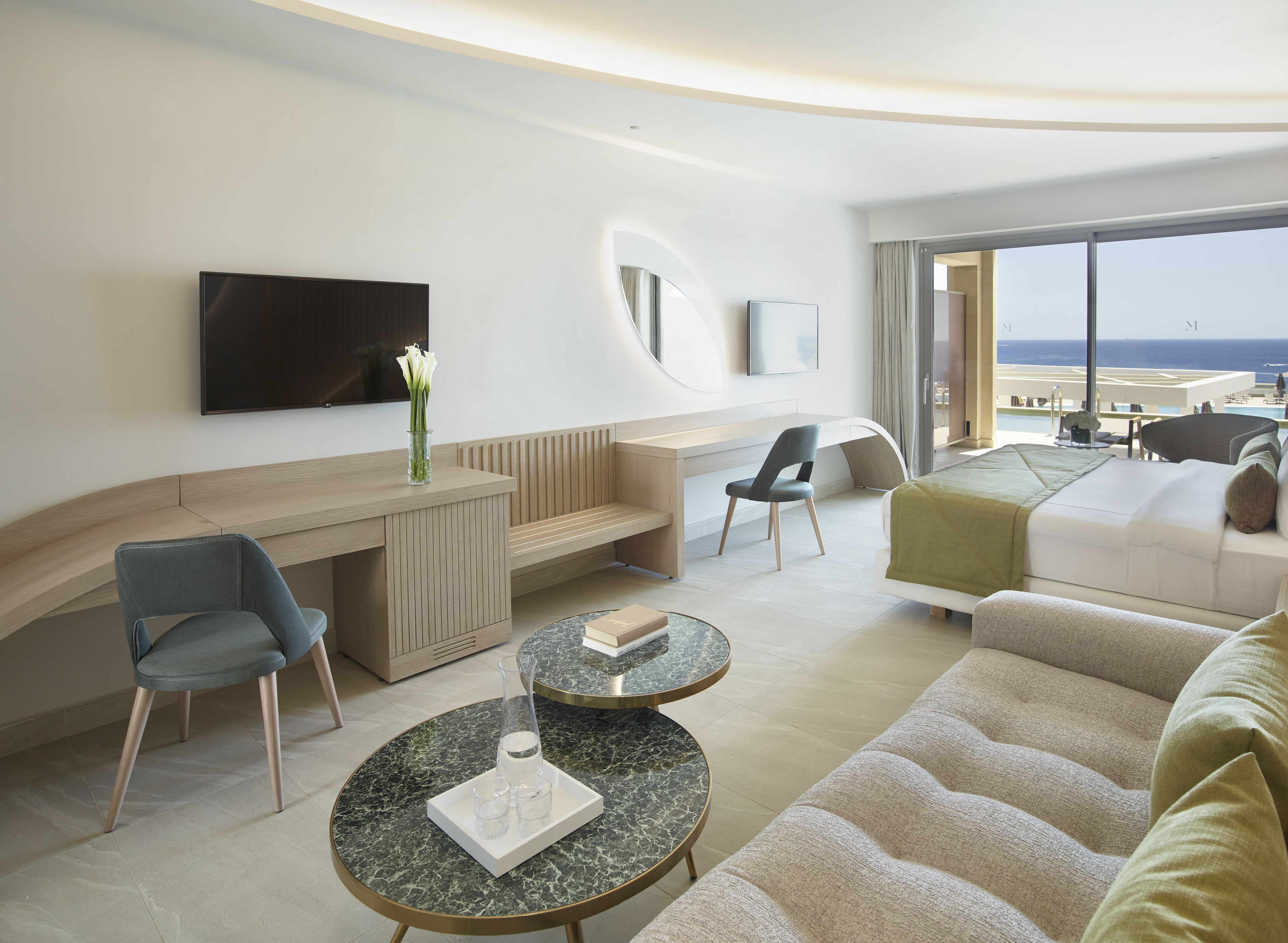 A modern hotel room with a curved desk, seating area, and a large window offering a view of the ocean.