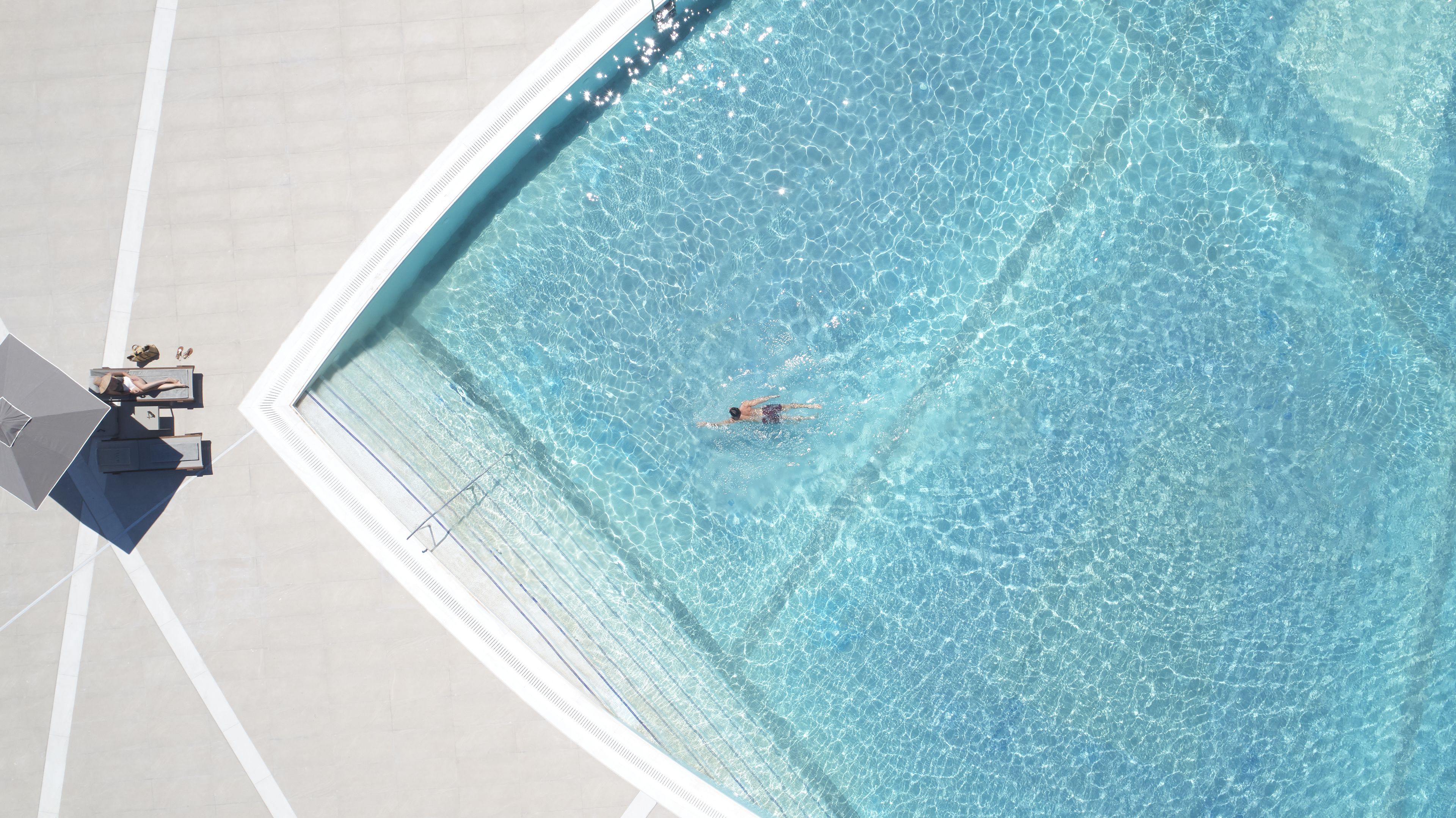 An aerial view of a swimming pool with clear water, a person swimming, and a lounge area with a table and chairs.