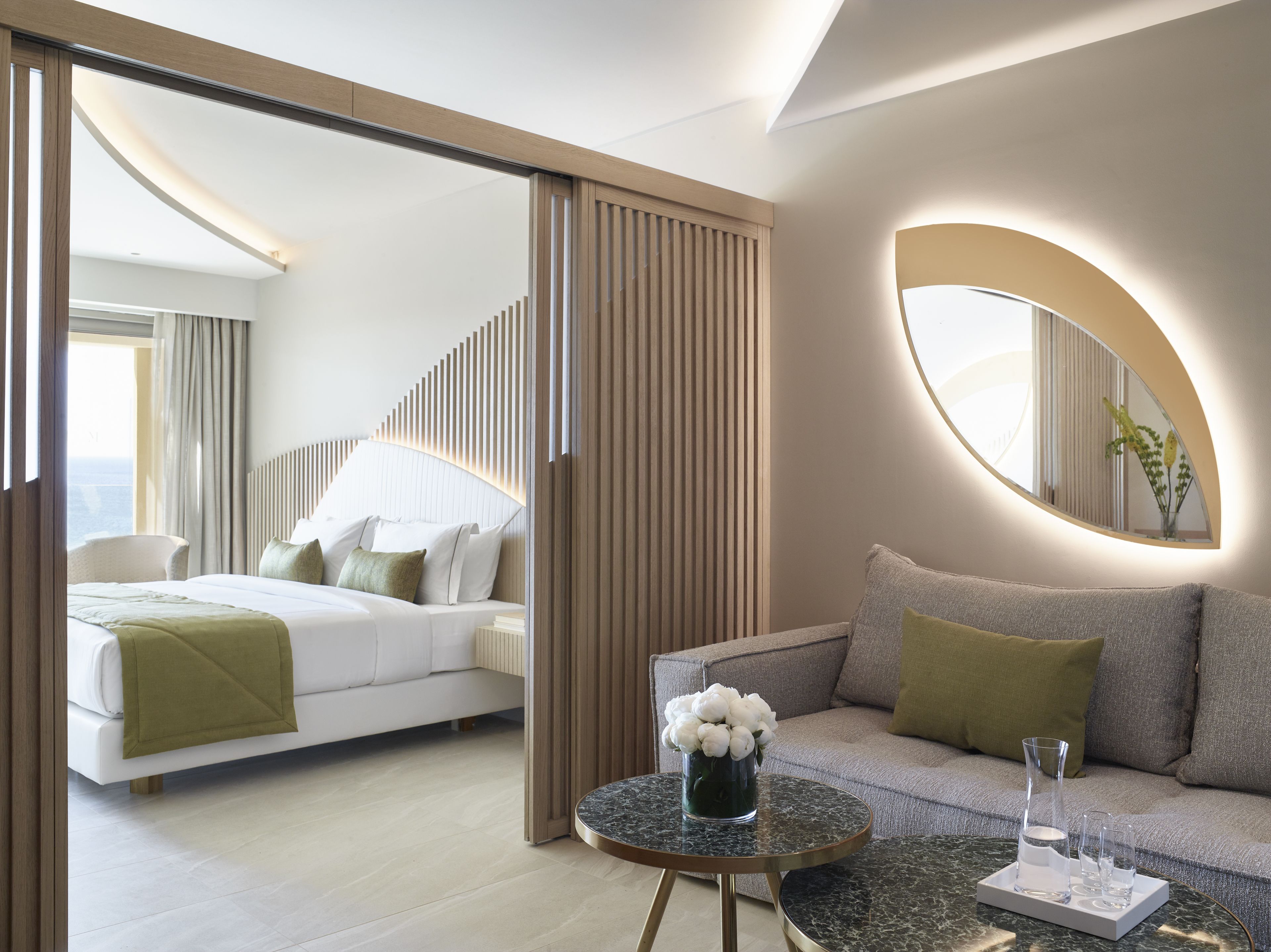 A modern hotel room with a bed, a sofa, a round table with a vase of flowers, and a large mirror.