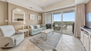 A living room with a sofa, coffee table, and a television on a cabinet. There is a balcony with a view of the landscape. - Waterfront Views | Boat-Friendly | Lazy River & Pools | On-site Dining (Orange Beach)