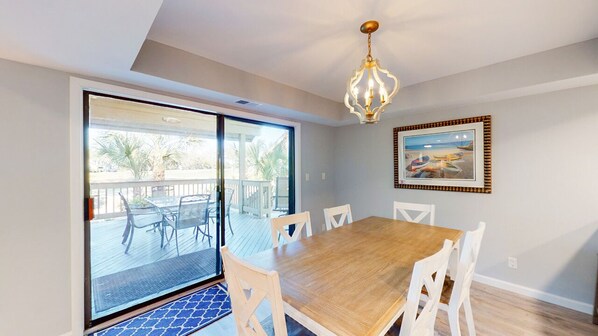 A dining area with a wooden table and white chairs, a chandelier, a large sliding glass door leading to a balcony, and a framed picture on the wall.
