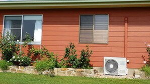 Une maison à l’extérieur rouge, avec deux fenêtres dotées de volets, un mur de soutènement en pierre et un climatiseur blanc.
