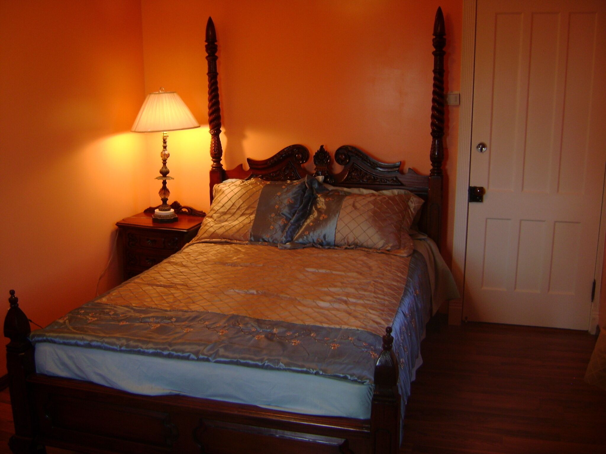 A four-poster bed with a wooden headboard, a bedside table with a lamp, and a door to the right.