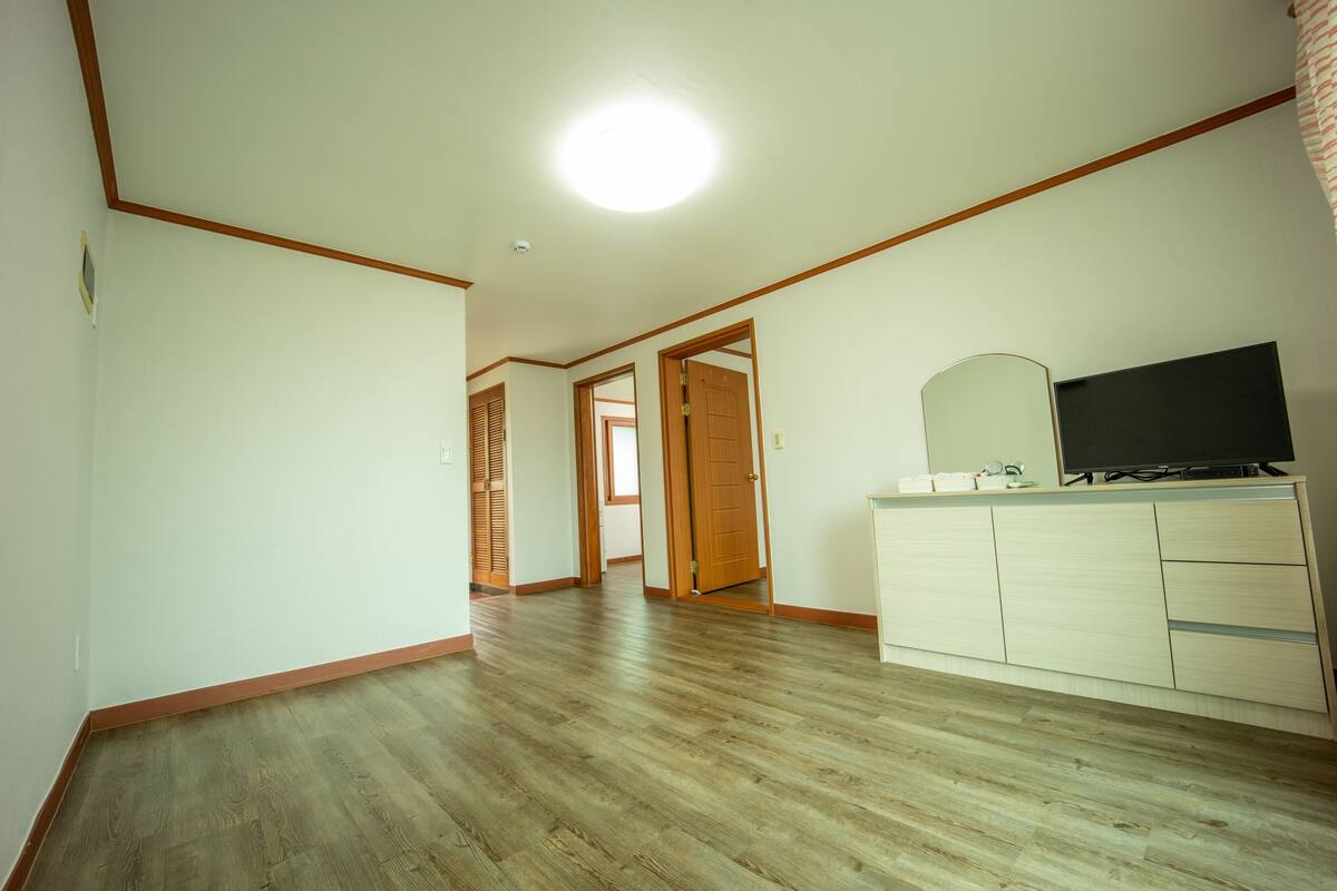 A room with wooden flooring, a white dresser with a mirror and television, and a door leading to another room.