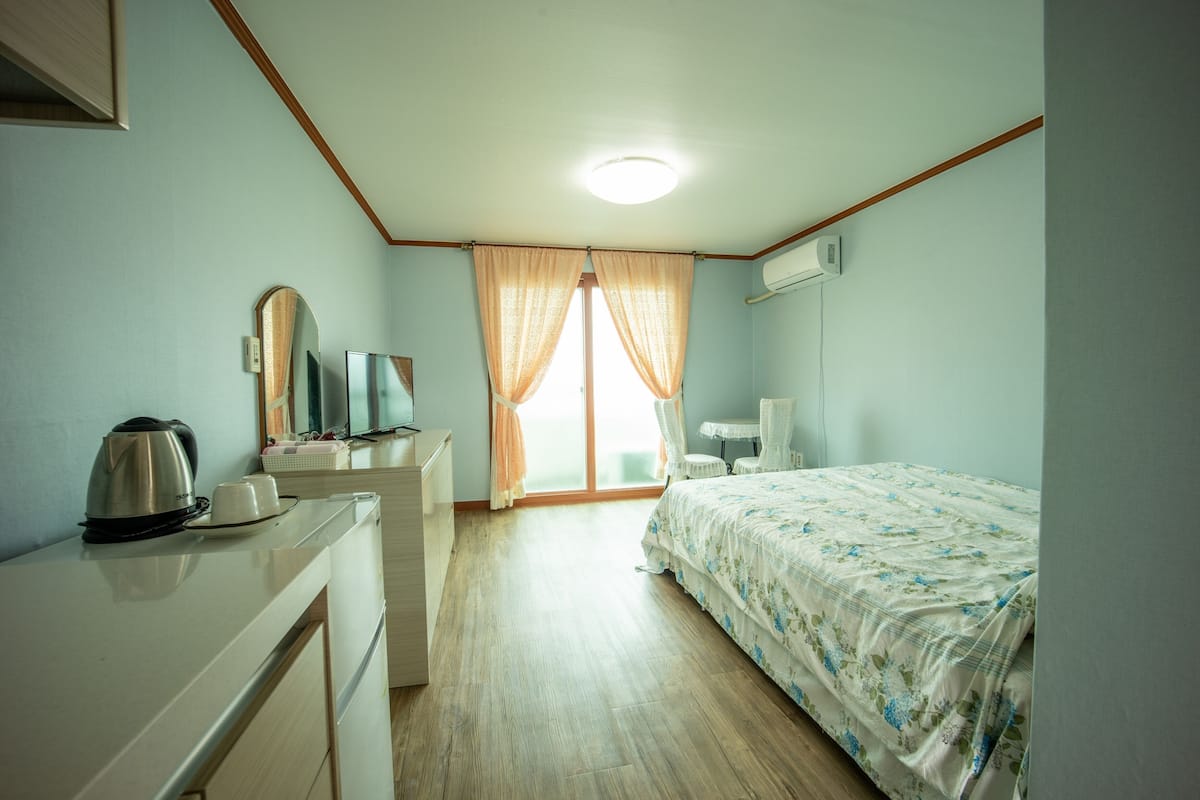 A hotel room with a bed, a desk with a kettle, a television, and a window with curtains.