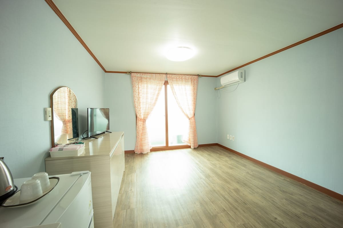A room with a wooden floor, a flat-screen TV, a mirror, a window with sheer curtains, and an air conditioning unit.