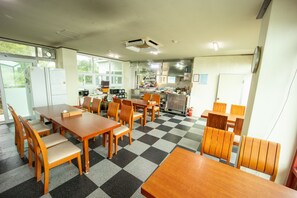 A dining area with checkered flooring, wooden tables and chairs, a refrigerator, and a kitchen in the background.