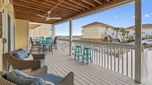 Une terrasse extérieure couverte, aménagée avec des meubles en osier, un ventilateur de plafond et offrant une vue sur les maisons de plage.