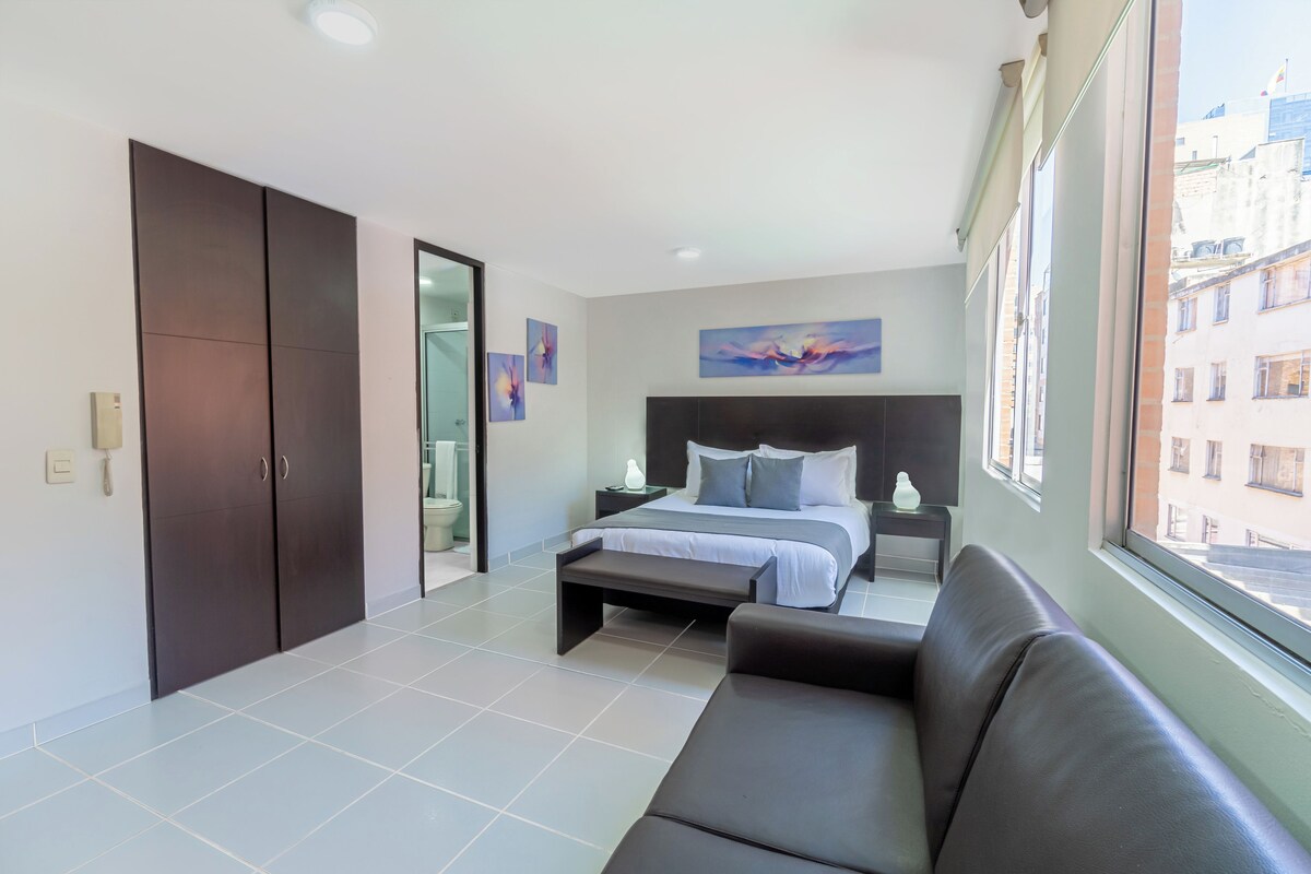 Un dormitorio moderno con cama, banco, sofá, armario y un ventanal grande.