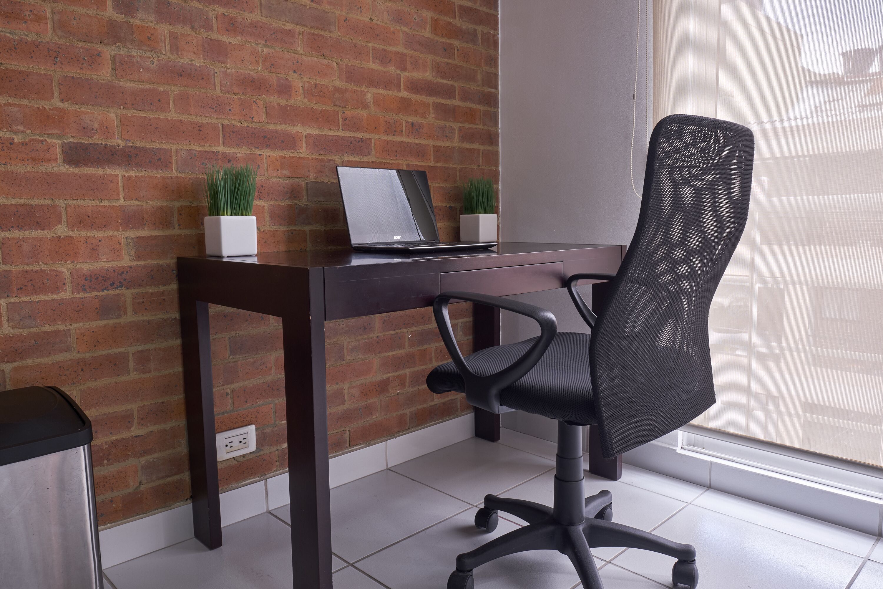 A workspace with a laptop on a desk, two potted plants, a black mesh office chair, and a brick wall.