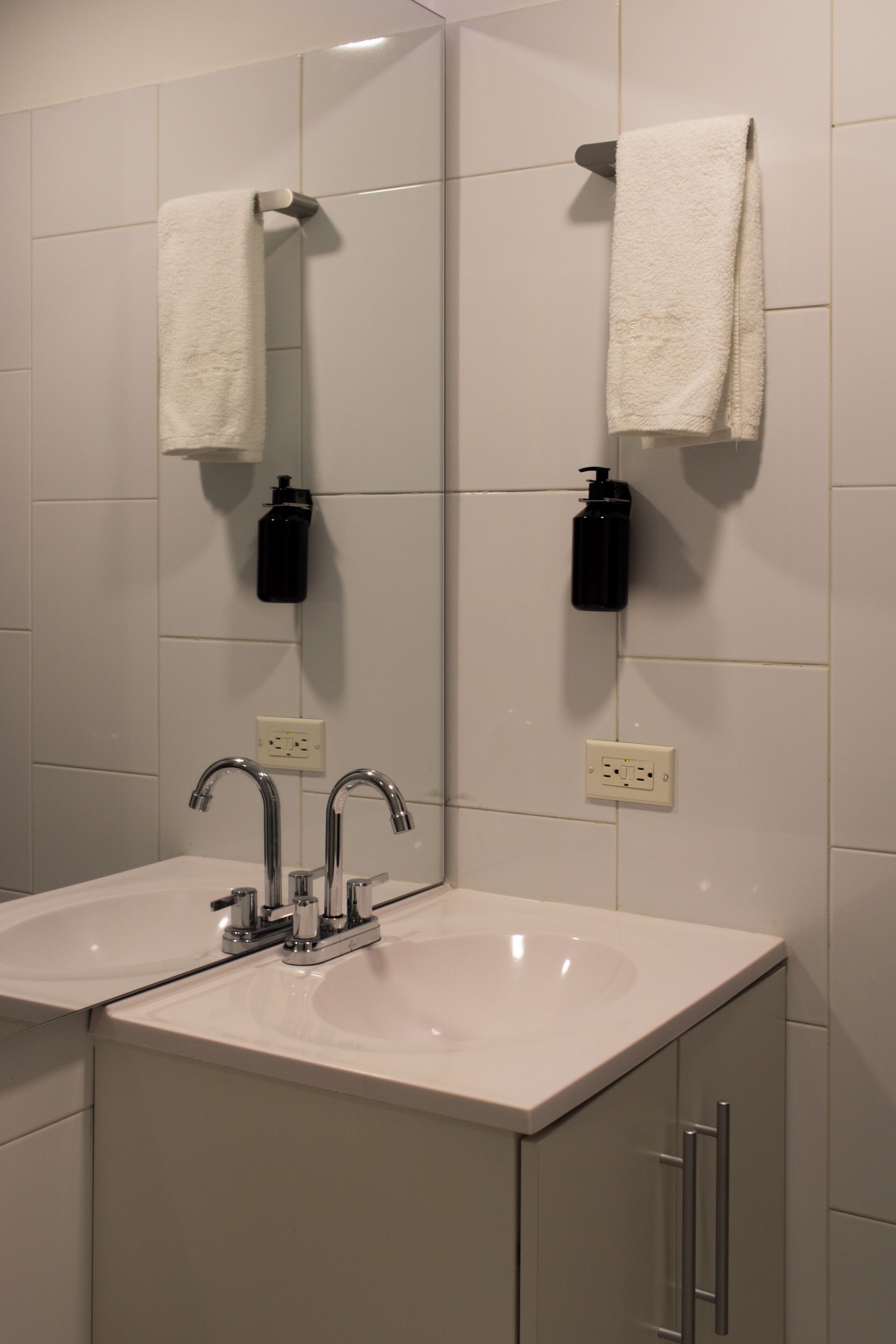 Un baño con un lavamanos blanco, dos llaves y un espejo. Hay dos dispensadores de jabón negros montados en la pared.