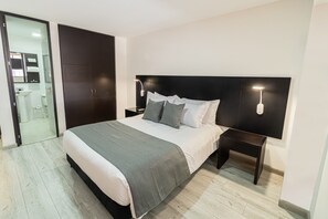 A modern bedroom with a large bed, dark headboard, bedside lamps, and a view into the bathroom.