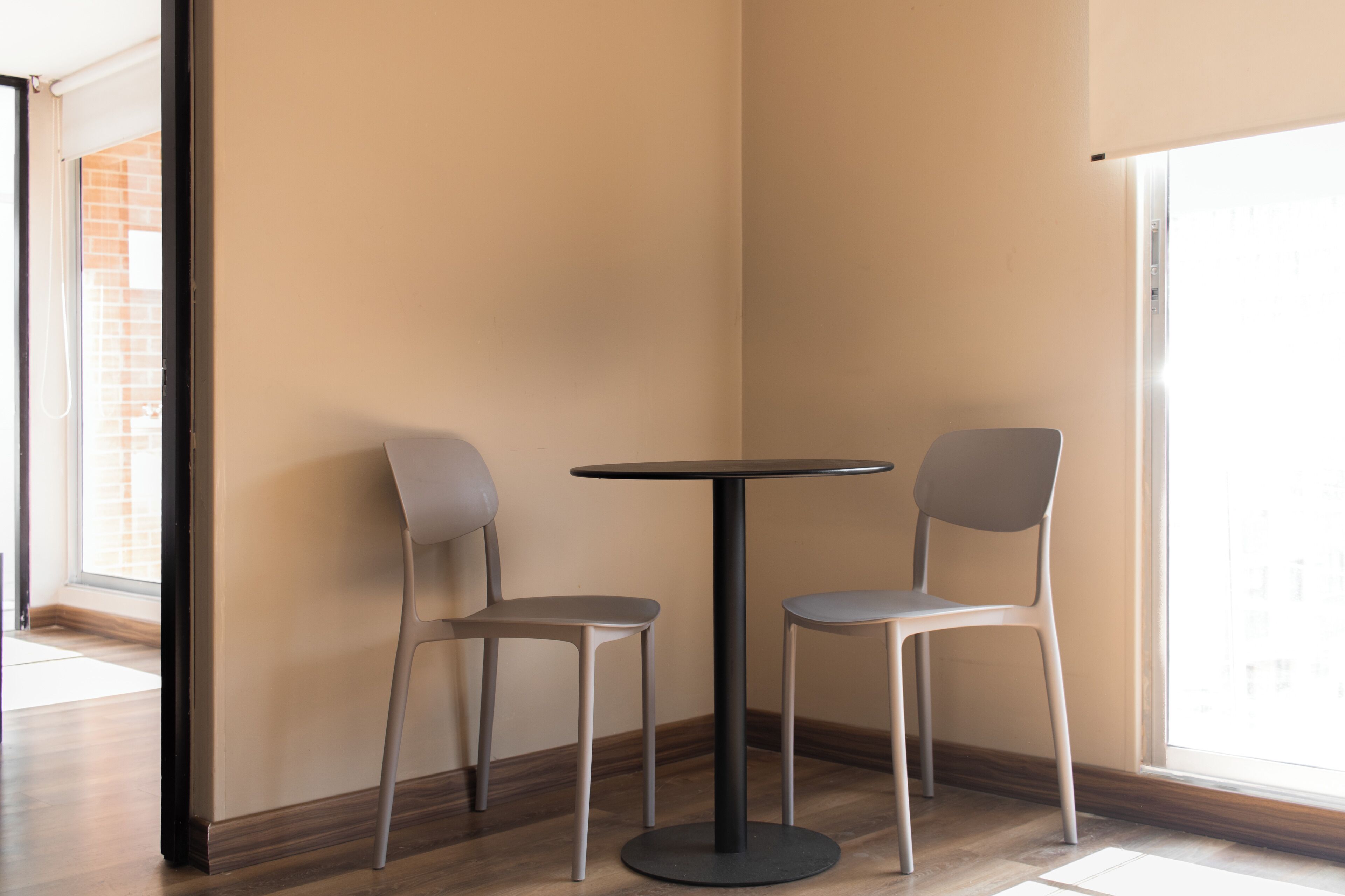 A corner room with a round table and two chairs, a window with a view of a brick building, and a wooden floor.